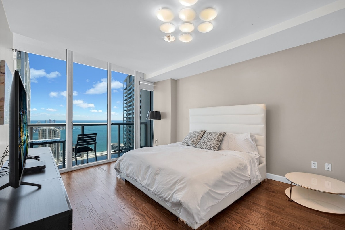 A spacious bedroom is highlighted by a king-size bed with a plush white headboard. Large windows offer views of Biscayne Bay, while a sleek television is positioned on a minimalistic black console. Natural light fills the room through the clear glass panels.