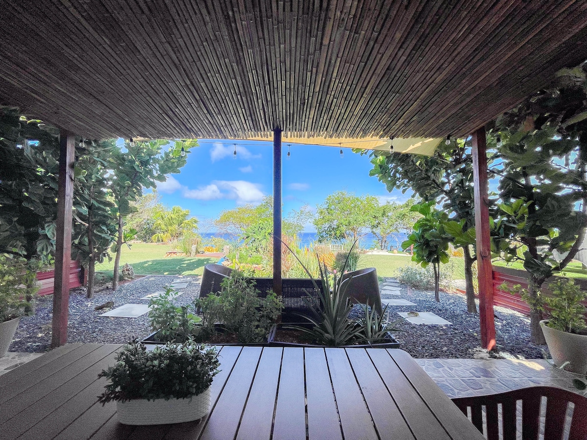 An outdoor seating area with a wooden table overlooks lush greenery and the ocean. Sunlight filters through the bamboo roof, creating a dappled effect. Tropical plants line the space, enhancing the connection to nature and providing a serene environment.
