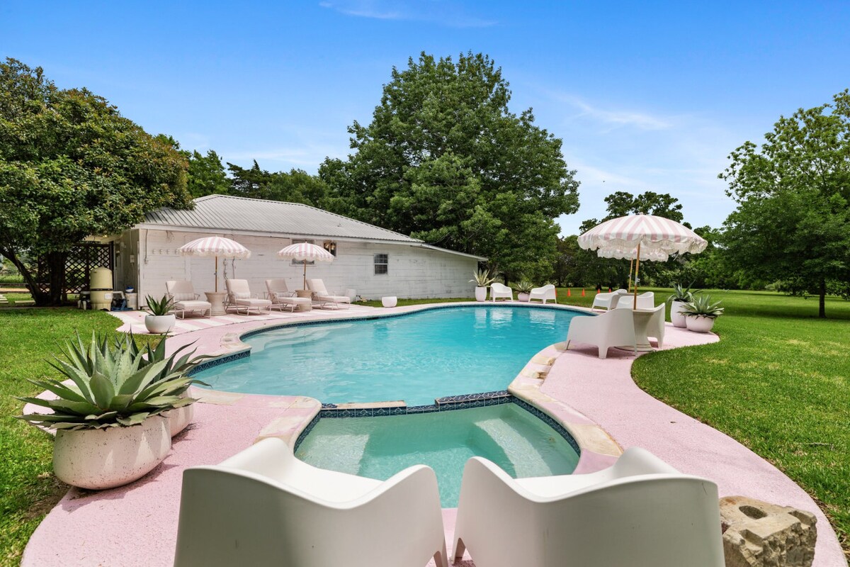 A serene pool area is featured, surrounded by a pink deck and lush greenery. Two striped umbrellas provide shade over white lounge chairs. The pool's inviting water reflects the sky, offering a refreshing spot for relaxation. The well-maintained lawn complements the tranquil setting.