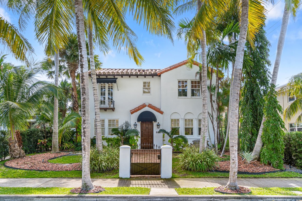 The exterior of the villa is showcased, featuring a charming two-story structure with a distinctive Spanish-style architecture. Lush palm trees and tropical landscaping frame the entrance, while a decorative wrought-iron gate leads to a welcoming pathway. The bright facade is complemented by vibrant greenery.