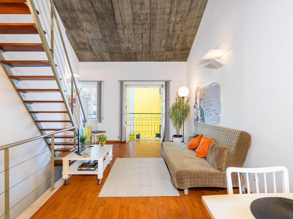 A modern living area features a spacious sofa adorned with orange cushions. Natural light illuminates the space through large glass doors that open to a balcony. A staircase with wooden steps leads to an upper level, complemented by white furniture and a neutral rug.