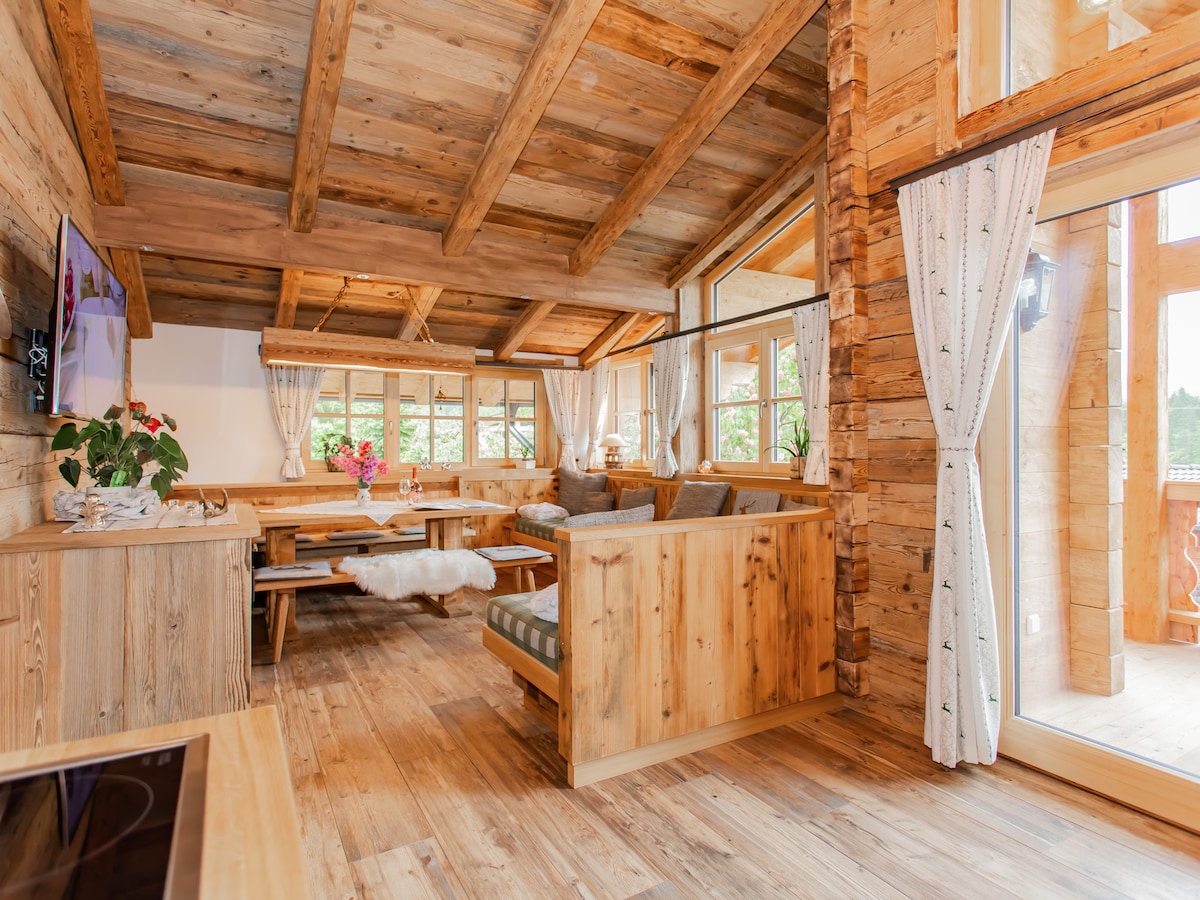 The open living and dining area features a wooden-beamed ceiling and ample natural light from large windows. A cozy seating arrangement is complemented by a dining table, creating a welcoming space for relaxation. Decorative plants add a touch of greenery to the earthy color palette.