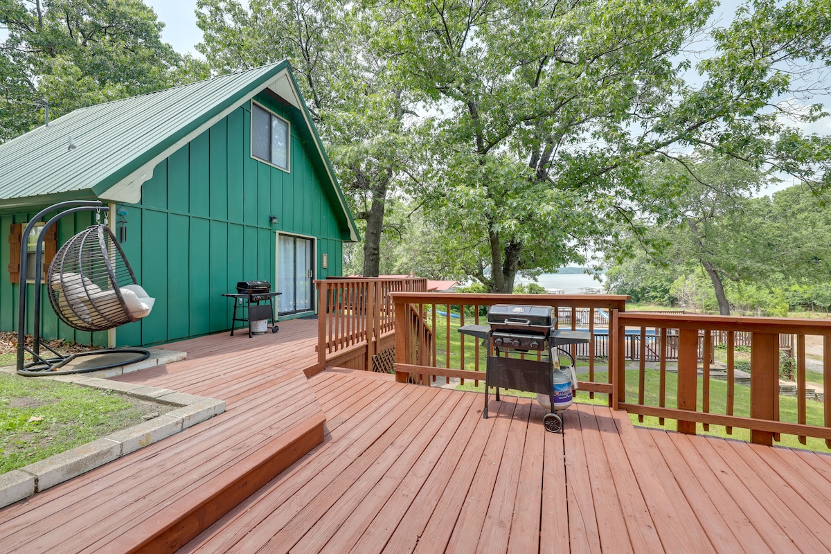 A spacious deck showcases multiple outdoor seating areas, including a hanging chair. A gas grill is positioned nearby, with a view of the surrounding greenery. The structure is highlighted by green siding and a sloped roof, complementing the natural landscape.