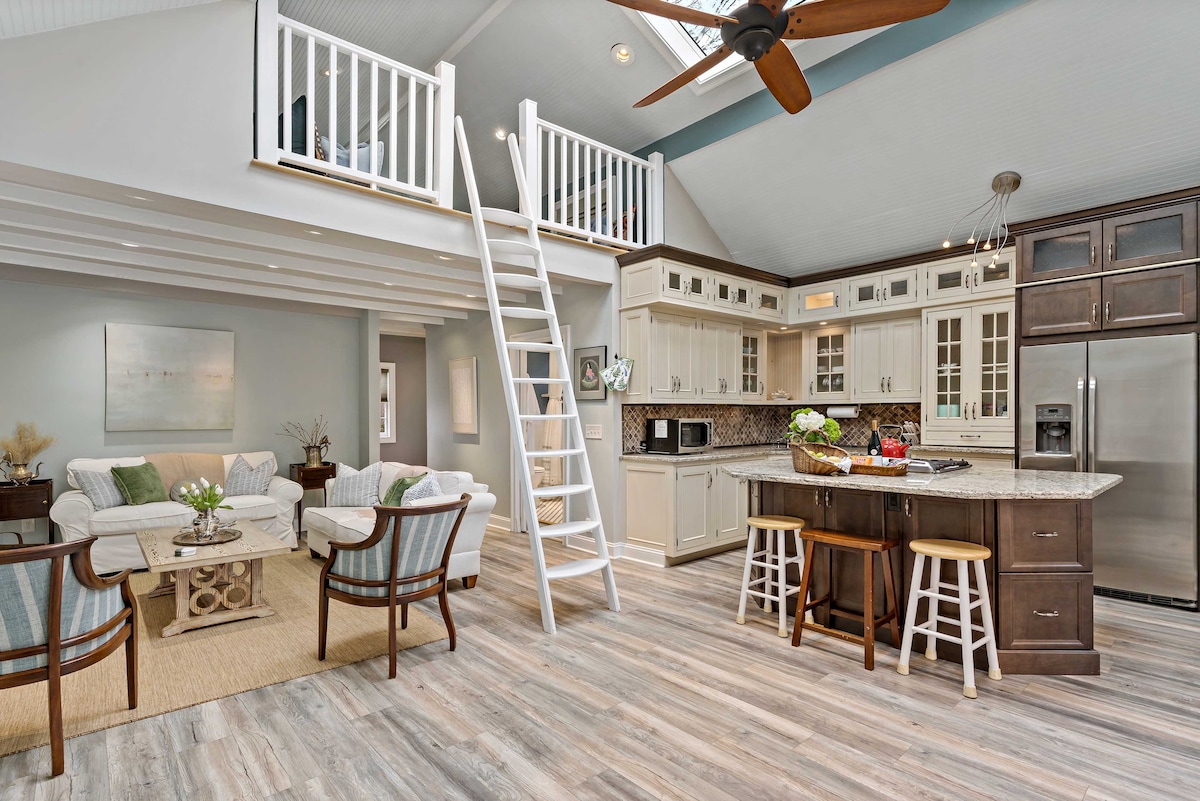 An open-plan living space showcases a modern kitchen with stainless steel appliances and an island with seating. A ladder leads to a loft area. Comfortable seating is arranged around a central table, and large windows fill the room with natural light.