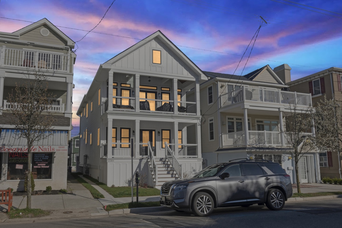 A two-story condo building is viewed from the street, showcasing a light exterior and multiple windows. The front steps lead to a welcoming porch. A parked SUV is visible in the foreground, with a backdrop of pastel-colored clouds in the evening sky.