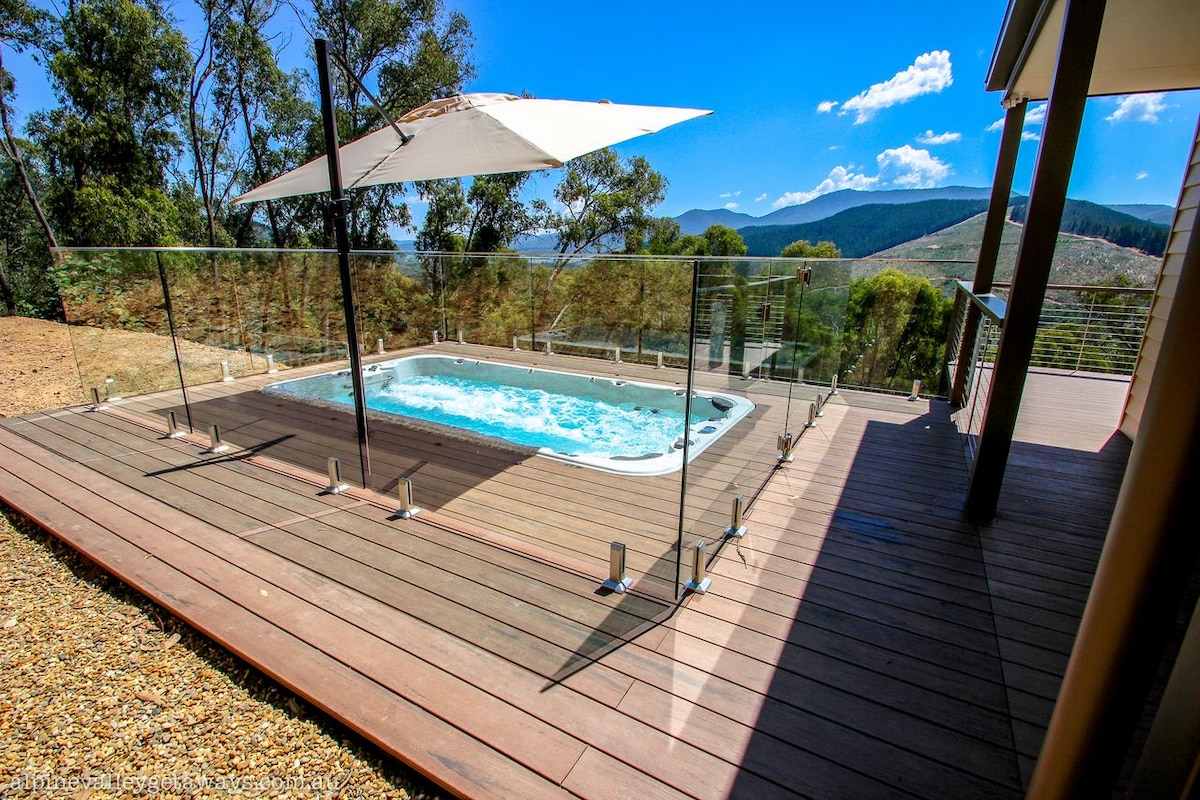 A climate-controlled pool-spa is featured on a wooden deck surrounded by natural scenery. Glass fencing enhances the view, while a large umbrella provides shade. The backdrop showcases mountains and a clear blue sky, creating a serene outdoor space for relaxation.