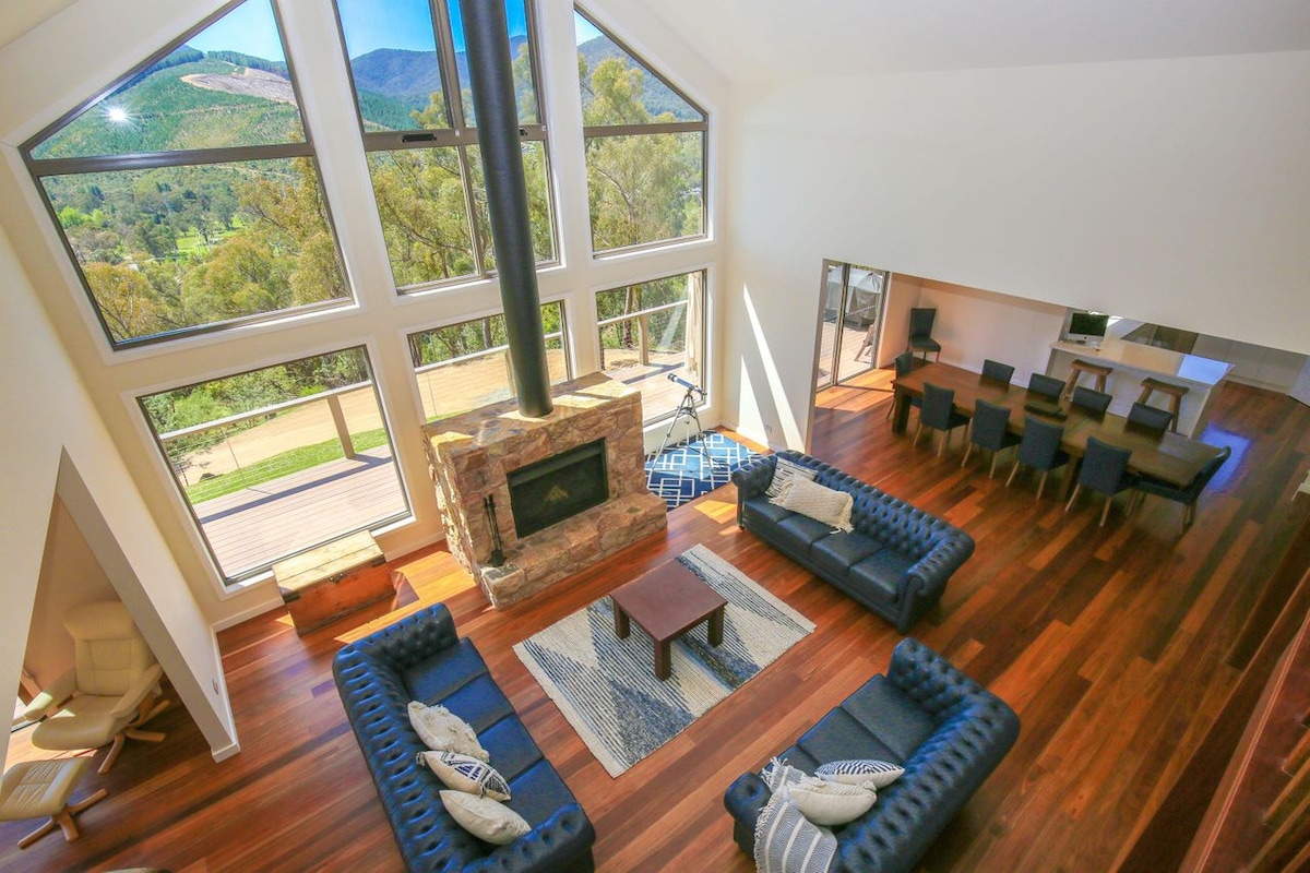 The spacious living area features large floor-to-ceiling windows, allowing natural light to fill the space and offering views of the surrounding mountains. A stone fireplace serves as a focal point, complemented by dark leather sofas arranged around a central coffee table, with a dining area visible in the background.
