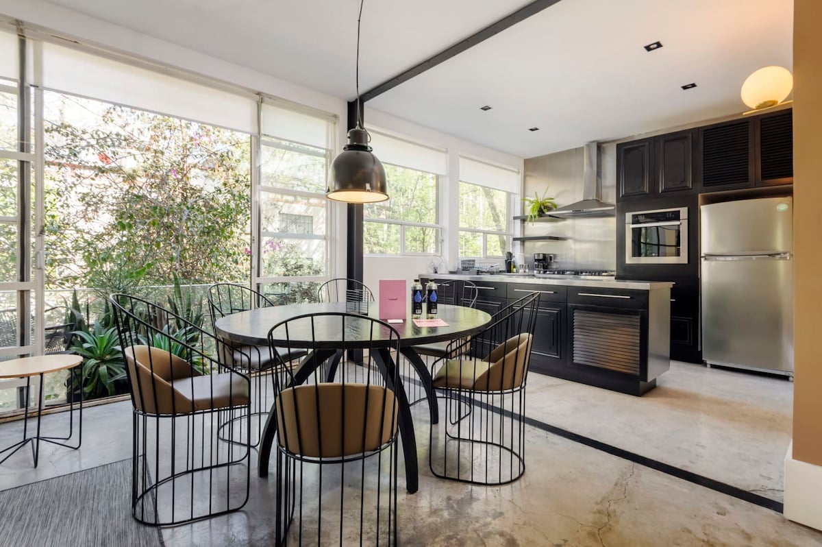 A modern kitchen and dining area features a round dining table surrounded by black wire chairs. Large windows provide ample natural light and a view of greenery outside. Sleek countertops and stainless steel appliances enhance the contemporary design of the space.
