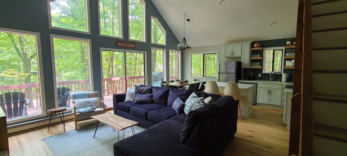An open living area features large windows that provide abundant natural light and views of the surrounding trees. A spacious sectional sofa and a wooden coffee table are arranged on a light area rug. The adjoining kitchen is equipped with modern appliances and a dining area for casual meals.