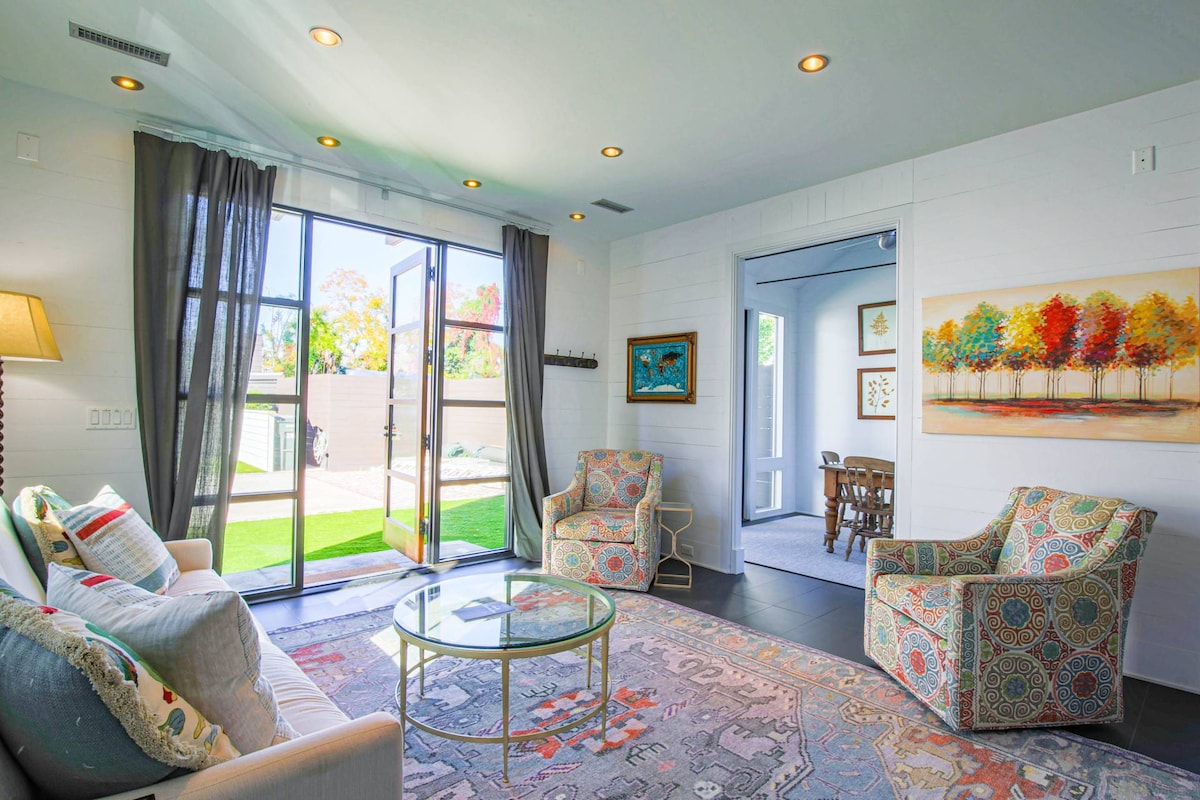 A bright living area is illuminated by natural light streaming through large glass doors that open to the private courtyard. Comfortable patterned chairs are positioned near a glass coffee table, complemented by a decorative area rug. A hallway leads to additional spaces within the home.