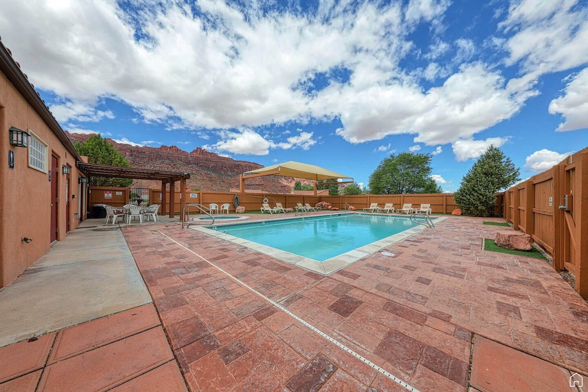 A spacious outdoor area features a seasonal swimming pool surrounded by a paved patio. Several lounge chairs are positioned around the pool, with shaded seating available under a canopy. The backdrop displays a clear blue sky with scattered clouds and distant rock formations.