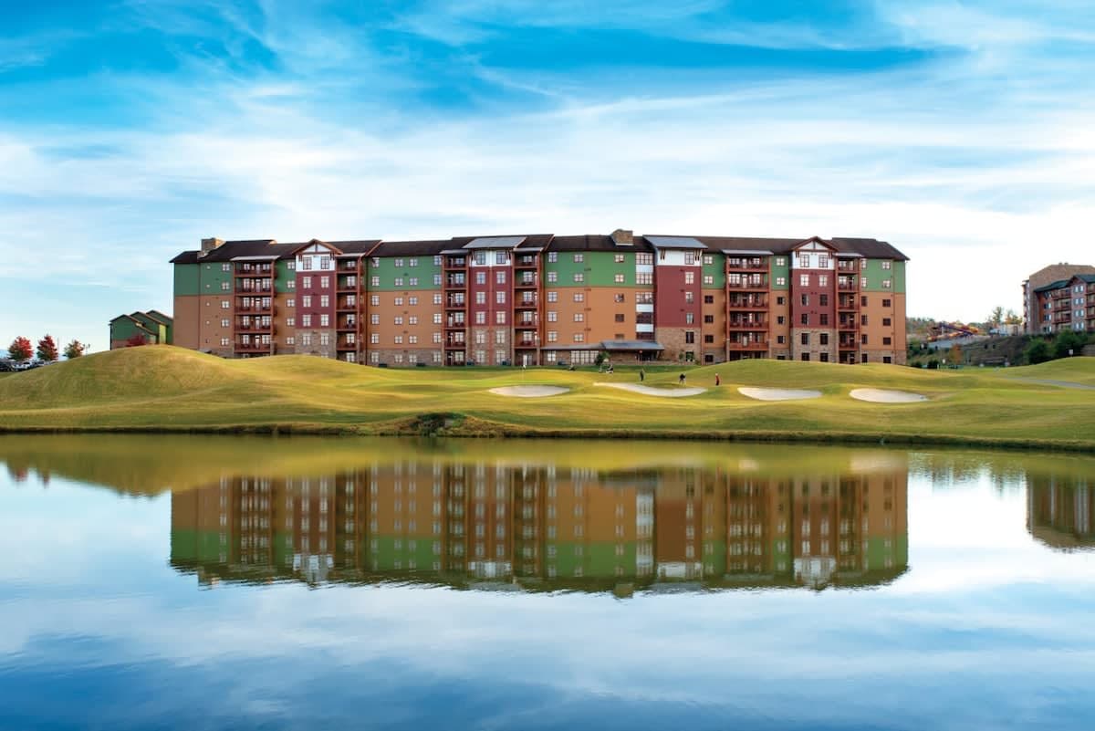 A large, multi-story lodge is set against a backdrop of rolling green hills and a serene lake. The building features a mix of red, green, and beige colors, reflecting beautifully on the calm water surface. A gentle sky with light clouds completes the scenic view.