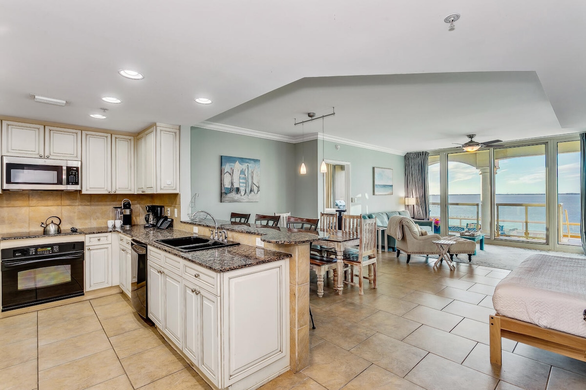 An open-concept kitchen and living area showcase modern appliances and a spacious granite countertop. Dining space includes a wooden table with matching chairs, while floor-to-ceiling windows reveal scenic views of the water. A cozy seating area with a sofa and beachside decor is presented.