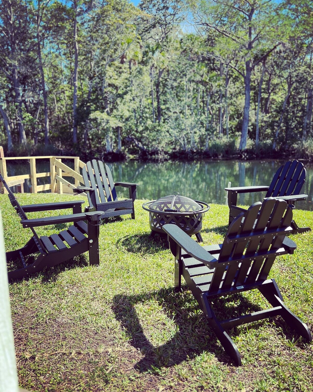 Four black Adirondack chairs are arranged around a fire pit on a grassy area beside a calm river. Lush green trees frame the scene, creating a serene outdoor space ideal for relaxation and gatherings.