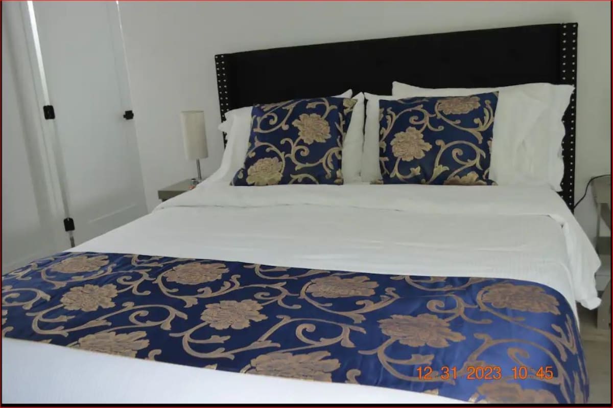 The master bedroom features a spacious bed adorned with decorative blue and gold accent pillows. A pair of bedside lamps provide subtle lighting on each side. A simple, modern style is complemented by the neutral color palette and soft linens.