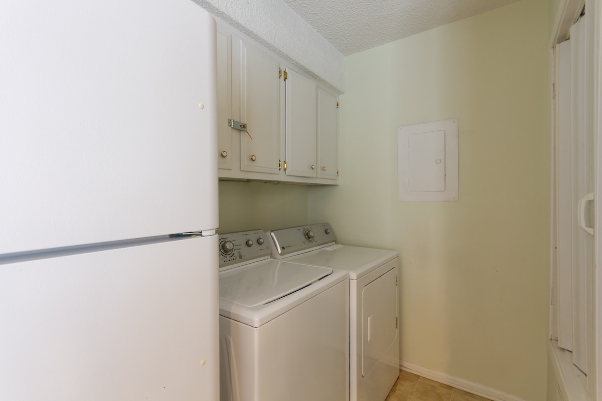 A dedicated laundry space is visible, featuring a stacked washer and dryer set beside a white refrigerator. Light-colored cabinetry and pale green walls create a clean and functional area, while a utility panel is discreetly mounted on the wall.