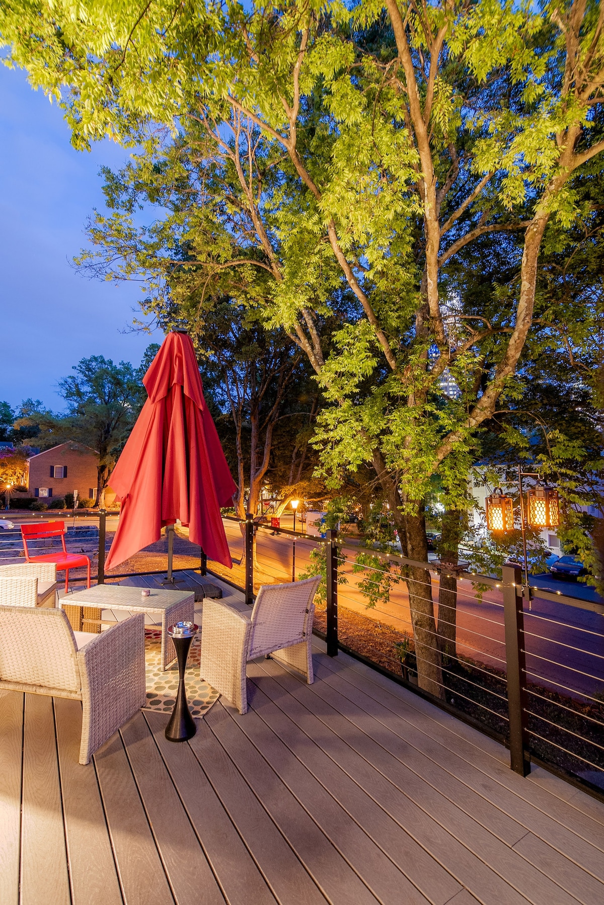 A spacious deck features two white wicker chairs beside a small table and a lighted umbrella. Tall trees provide shade, while nearby streetlights cast a warm glow. The surrounding neighborhood adds a tranquil backdrop to this inviting outdoor space.