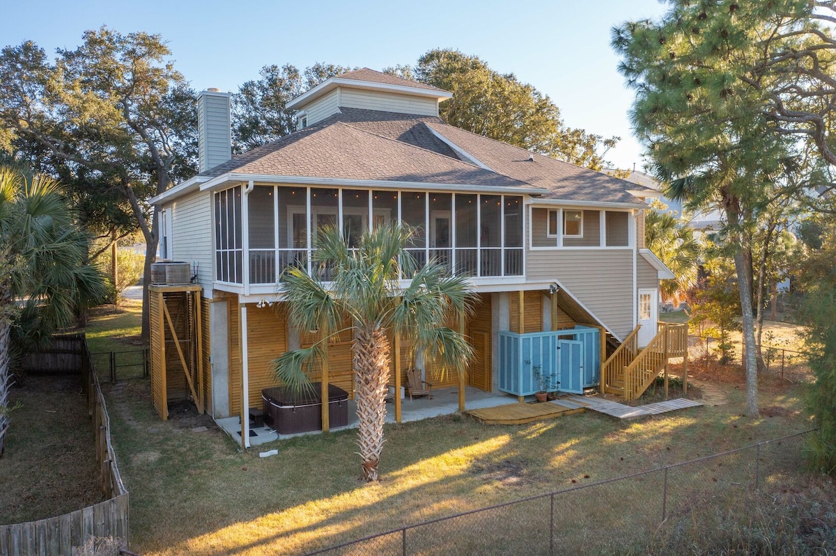 The exterior of a two-story home is showcased, featuring a spacious screened porch and a hot tub on the lower deck. Tall palm trees are visible in the yard, which is enclosed by a wooden fence. The natural surroundings include grassy areas and nearby trees.