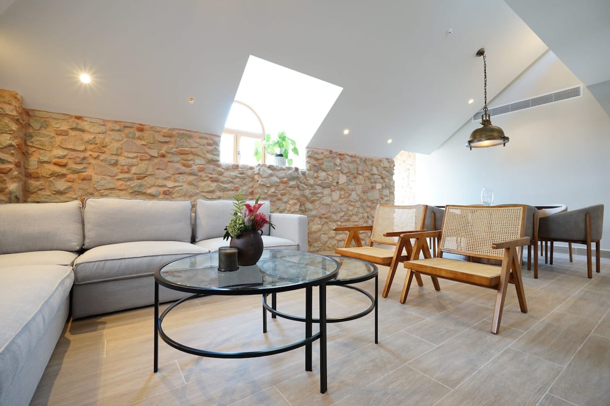 A spacious living area features a light-colored sectional sofa arranged around a round glass coffee table. Natural stone walls complement the modern decor. A set of two wooden chairs is positioned nearby, and windows allow light to enter, enhancing the overall brightness of the space.