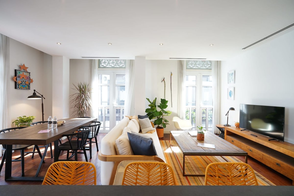 A spacious living area features a light-colored sofa and a wooden coffee table in front of a modern entertainment unit. Large windows allow ample natural light, and a dining table is set for six. Various plants add a touch of greenery to the inviting space.