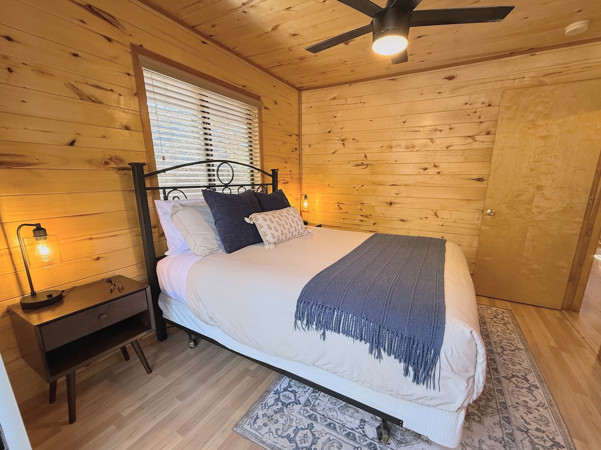 Acogedor dormitorio con paneles de madera con cama tamaño queen, ventilador moderno y lámparas cálidas.