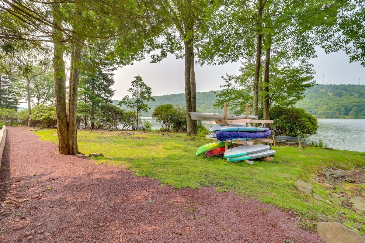 A grassy area bordered by trees offers various colored kayaks stacked for easy access. The water is visible in the background, with a gentle landscape of hills beyond, creating a serene outdoor space for recreation.