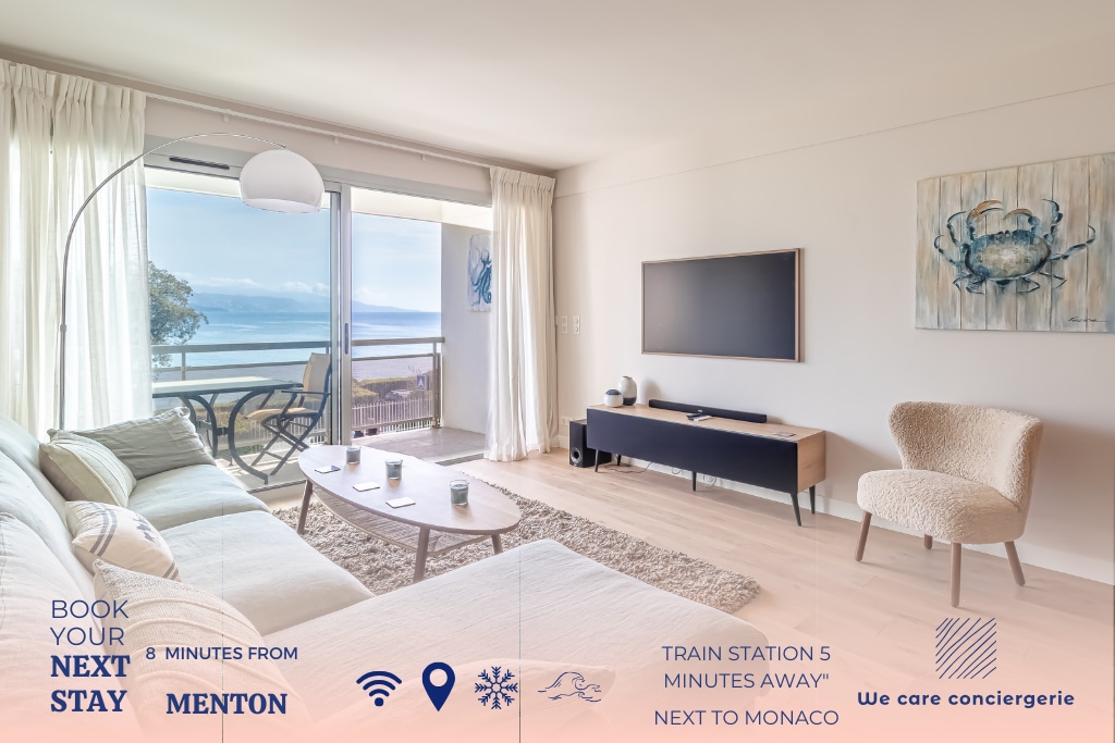 A bright living room is displayed, featuring a large pastel sofa and a television unit. Floor-to-ceiling windows provide a captivating sea view. Natural light fills the space, emphasizing the soft tones of the decor. A modern, relaxed atmosphere is created in this inviting area.