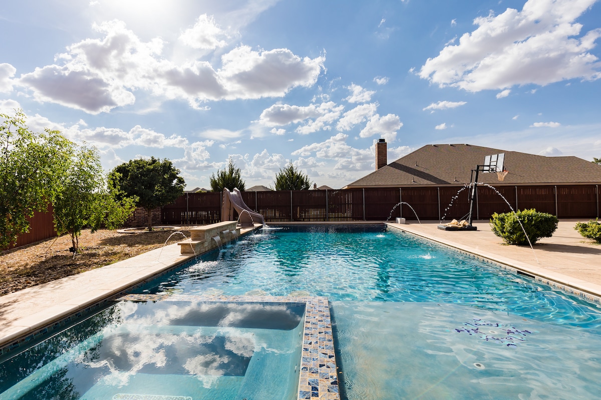 The spacious pool area features a large swimming pool with a slide and deck jets. A hot tub is positioned nearby, and lush landscaping surrounds the space. The backdrop includes a residential structure and a basketball hoop, under a bright sky with scattered clouds.