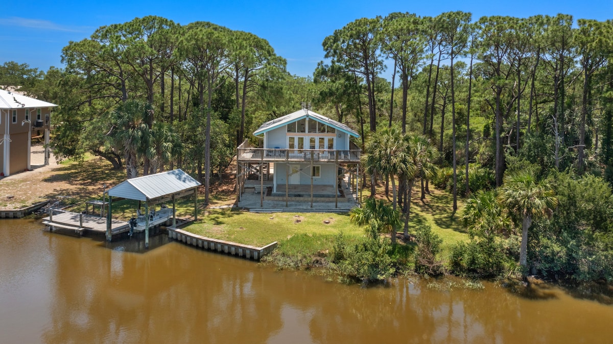The waterfront stilt home features a spacious outdoor deck and is surrounded by lush greenery, including tall pines and palm trees. A private dock is visible alongside the tranquil waters, providing a serene atmosphere in a natural setting.