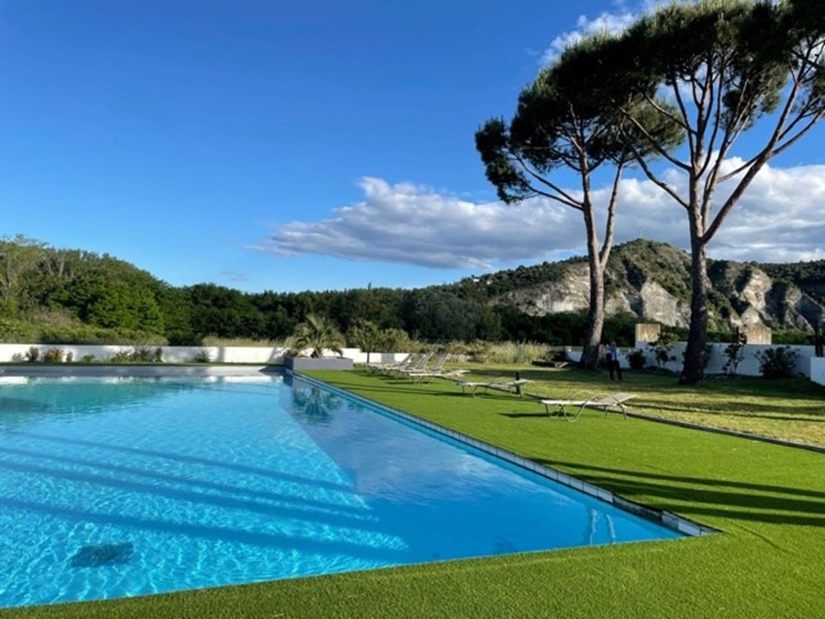 A large swimming pool is surrounded by a well-manicured grassy area. Lounge chairs are positioned around the pool, under the shade of tall trees. Lush greenery and hillside can be seen in the background, offering a relaxing outdoor environment.