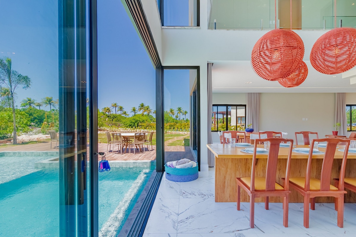 A dining area with a large wooden table and orange-accented chairs is visible alongside expansive glass doors, offering a view of the outdoor space that includes a swimming pool and landscaped gardens. Brightly colored pendant lights hang above the table.