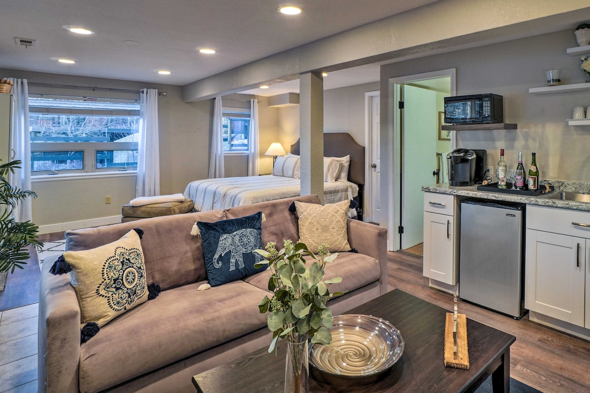 The open living area features a comfortable sofa with decorative pillows, a coffee table, and a kitchenette equipped with a refrigerator and microwave. Large windows allow natural light to brighten the space, which includes a king bed in the background and a separate entrance to the bathroom.