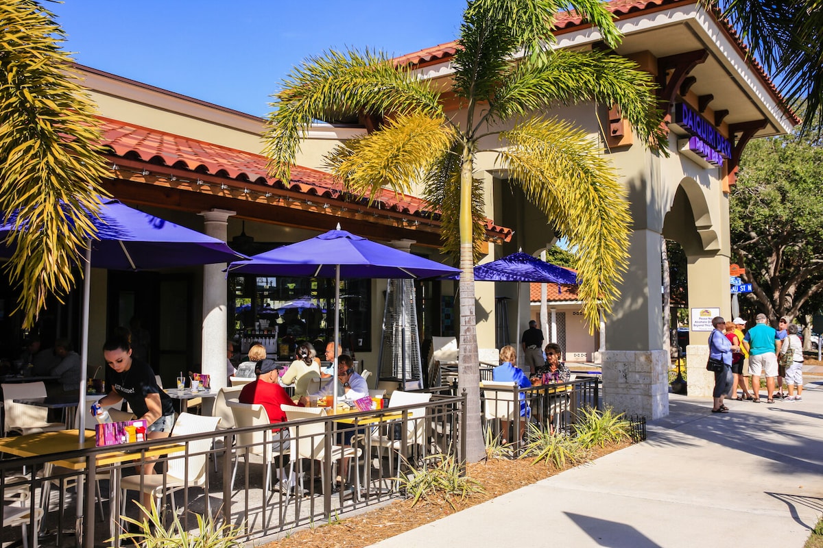 A vibrant outdoor dining area is shown with multiple tables under large umbrellas. Patrons are seated, enjoying their meals in a lively atmosphere. Decorative palm trees line the walkway, enhancing the tropical feel of the space. The building features a warm color palette with a tiled roof.