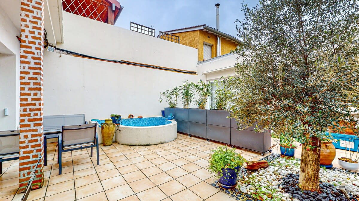 A spacious patio is presented, featuring a small round basin filled with blue water. Surrounding the basin, decorative stones and carefully arranged plants create a serene environment. An outdoor dining set with chairs is visible, along with a wall providing privacy and a touch of greenery from nearby trees.