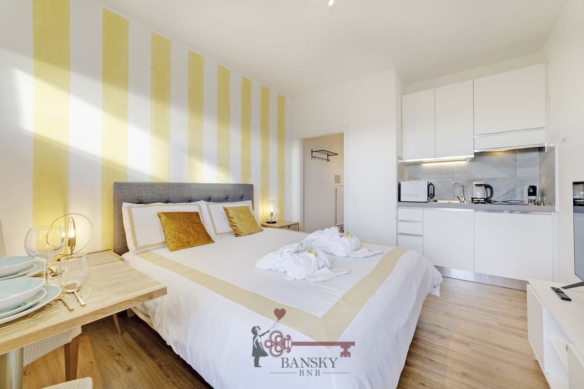 A spacious bedroom is highlighted by soft yellow striped walls, creating a bright atmosphere. The large bed is dressed with white linens and accented by golden cushions. A modern kitchenette is visible in the background, equipped with essential appliances for convenience.