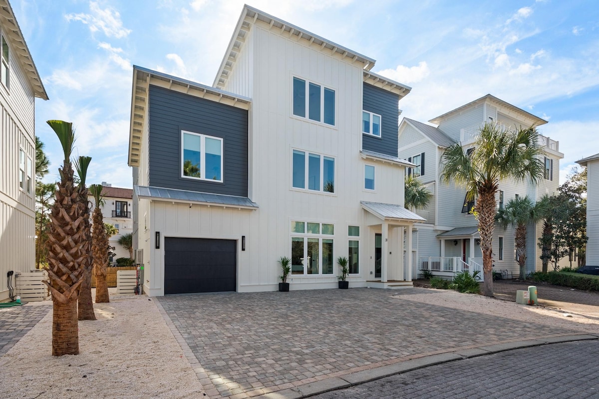 A modern three-story vacation rental home showcases a light exterior with white and gray paneling. Large windows line the front, allowing natural light to flood the interior. A spacious driveway is paved with bricks, featuring palm trees and manicured landscaping on either side.