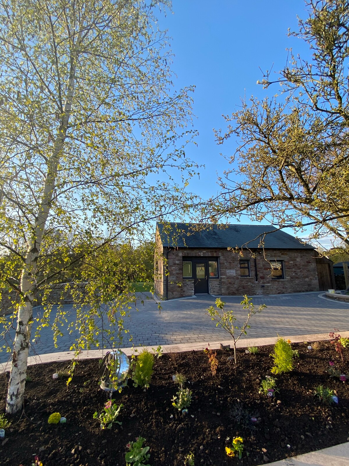 The exterior of a renovated barn conversion is shown, featuring a stone façade and a dark roof under a clear blue sky. Lush greenery surrounds the entrance, with new plants and flowering shrubs adding a touch of color to the well-maintained landscaping.