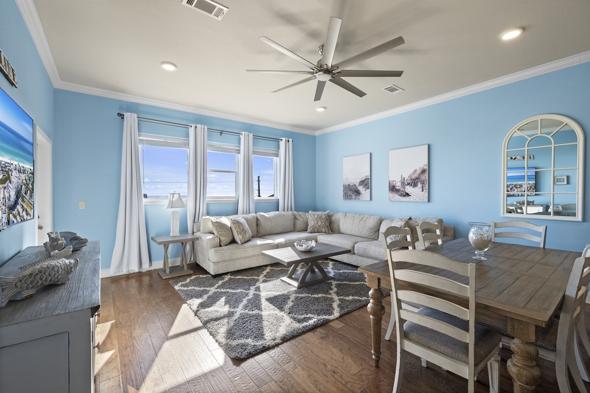 A spacious living area features light blue walls and hardwood flooring. A comfortable sectional sofa is positioned in front of a mounted flat-screen TV. A wooden dining table accommodates six, while large windows provide natural light and a view of the surroundings. Neutral decor enhances the modern feel.