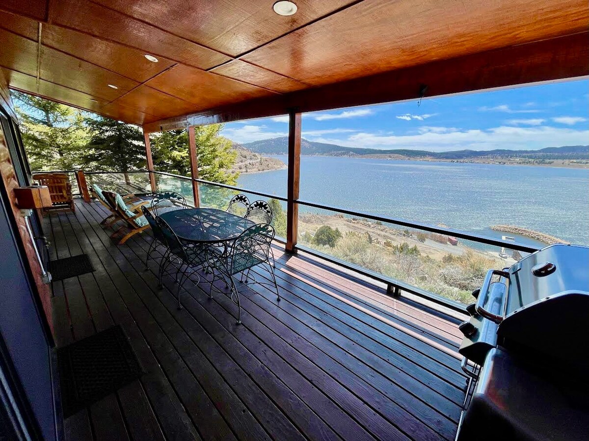 A covered deck offers outdoor seating with views of Panguitch Lake. A dining table is surrounded by chairs, and a propane grill is positioned nearby. Natural light filters through the railing, enhancing the serene atmosphere of the lakeside setting.