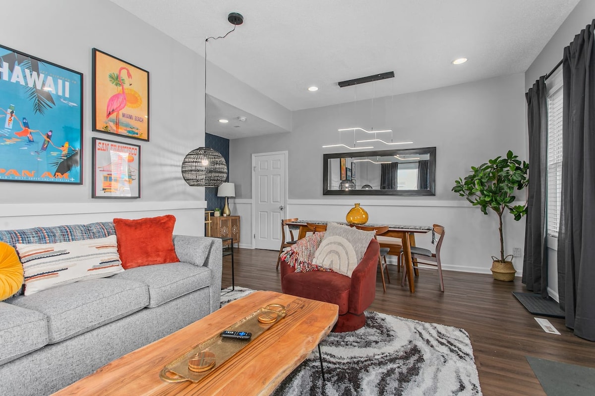 A stylish living area features a gray sofa adorned with colorful cushions and a patterned area rug. A wooden coffee table holds a remote, while elegant light fixtures accentuate the space. A dining table and decorative plants are visible, creating a cohesive and inviting atmosphere.