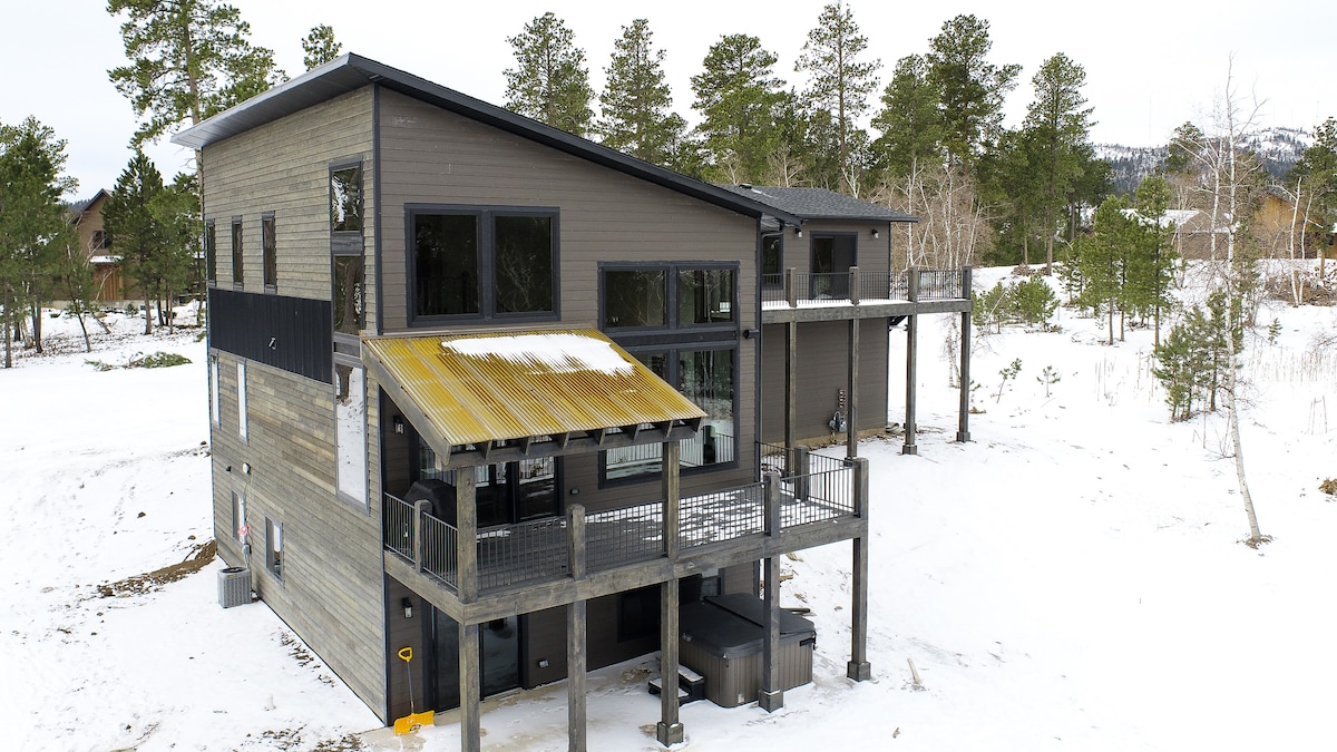 A contemporary cabin structure is showcased in a snowy landscape. The building features large glass windows and a covered deck, with a gold-colored awning extending out. The surrounding area is dotted with evergreen trees, providing a natural backdrop.