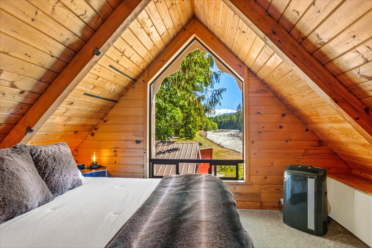 A cozy loft bedroom features an A-frame ceiling with wooden paneling. Large windows provide views of the river and surrounding trees, while a spacious bed is dressed with plush linens. Nearby, a small heater stands ready for warmth.