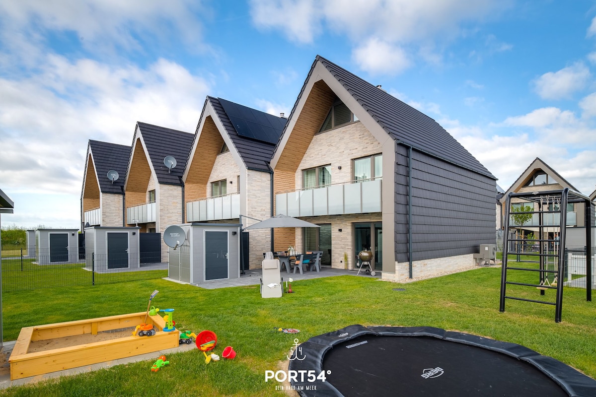 A modern, two-story vacation home features a mix of stone and dark siding. The exterior includes a covered terrace, a sandbox with toys, a trampoline, and a swing set in the yard, bordered by well-maintained green grass and a clear sky.