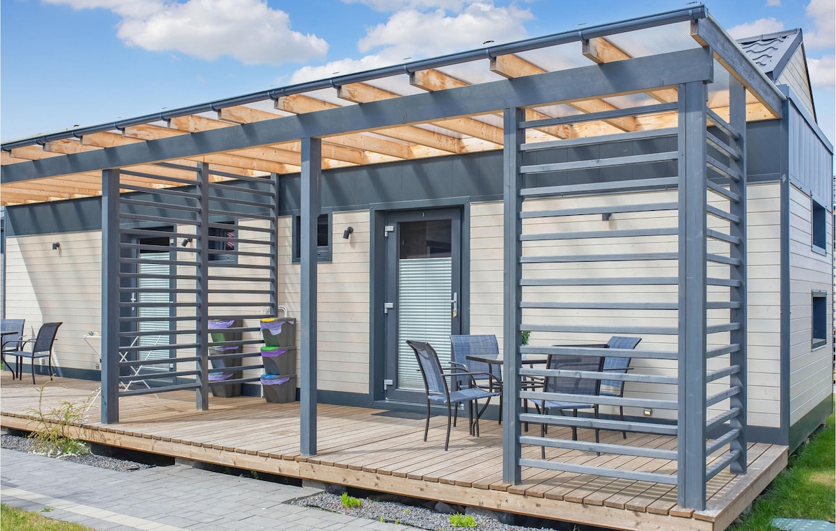 A modern vacation home is displayed with a spacious wooden deck, featuring a covered seating area with several chairs and tables. Vertical wooden slats create an inviting entrance, while colorful items hint at fun activities nearby.