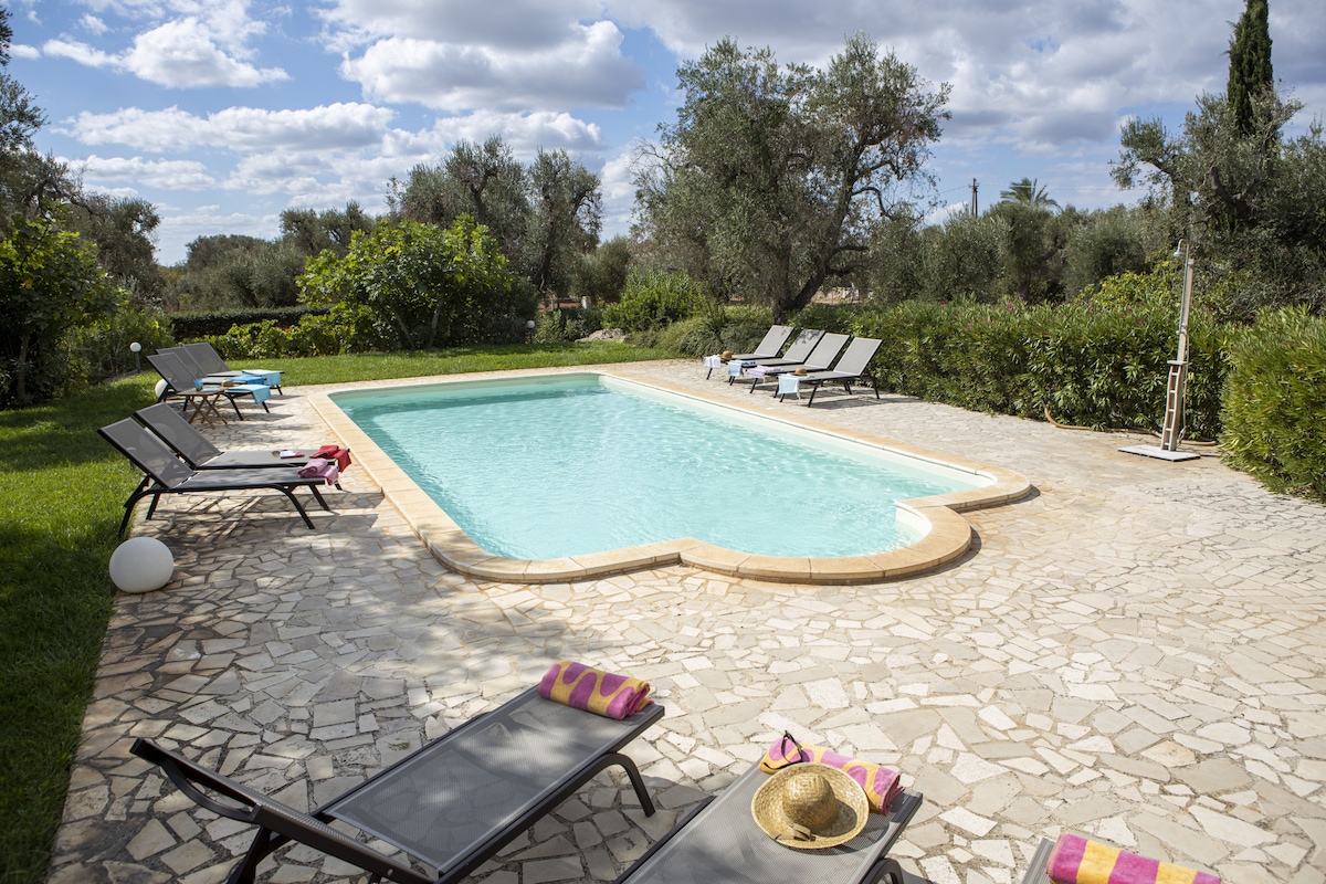 A tranquil pool area is surrounded by lush greenery, featuring a light blue pool set on a stone-tiled patio. Several lounge chairs are positioned around the pool, adorned with bright towels. A sun hat rests on one of the chairs, inviting relaxation.