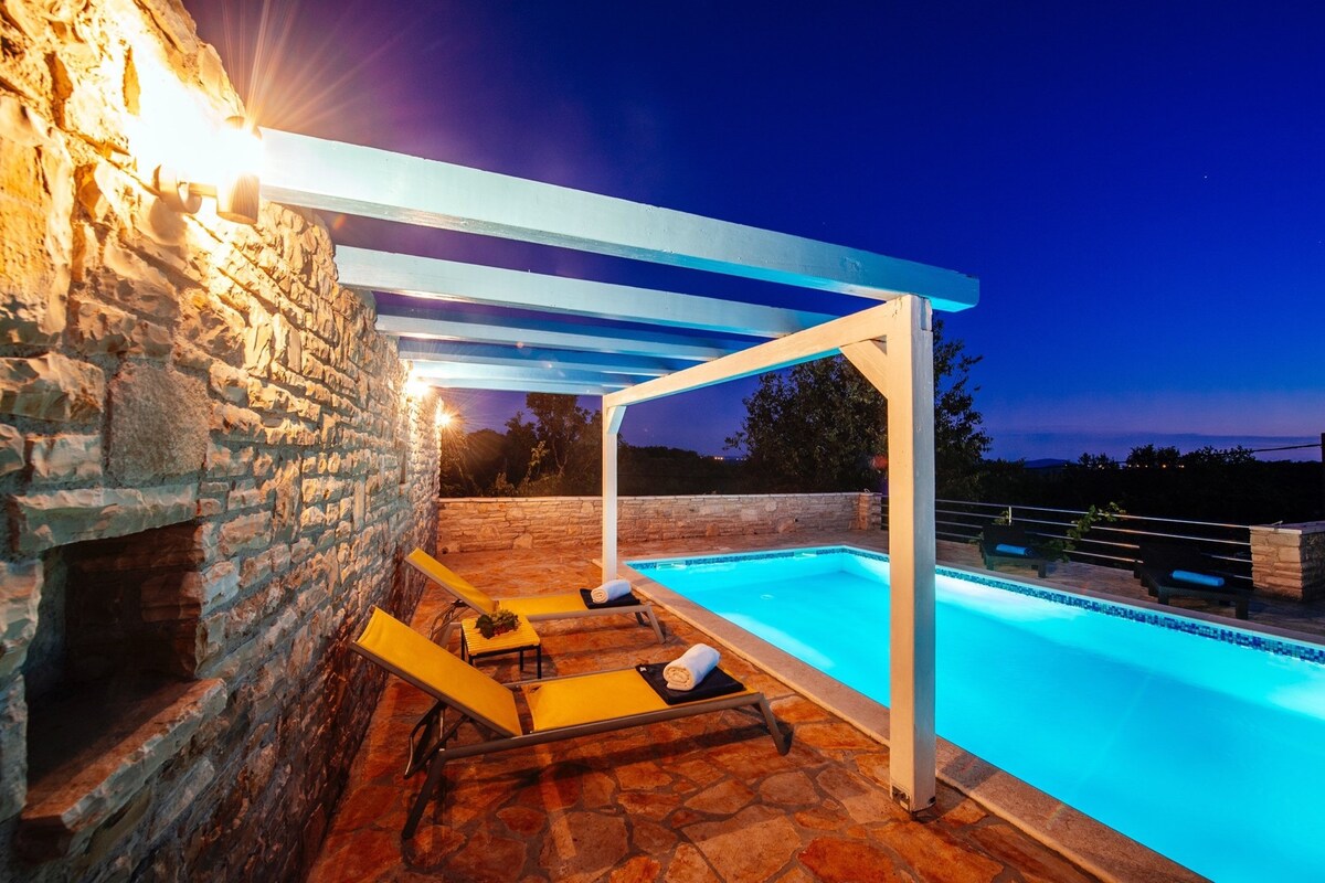 The outdoor space features a private pool under a vibrant blue sky, framed by a covered terrace supported by white beams. Stone walls and warm lighting create a cozy ambiance. Loungers are arranged alongside the pool, inviting relaxation within the serene surroundings.