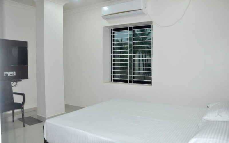 A spacious room featuring a large bed with crisp white linens. A wall-mounted television is positioned near a black chair, and a window with blinds allows natural light to enter. The clean, minimalist design enhances the serene atmosphere.