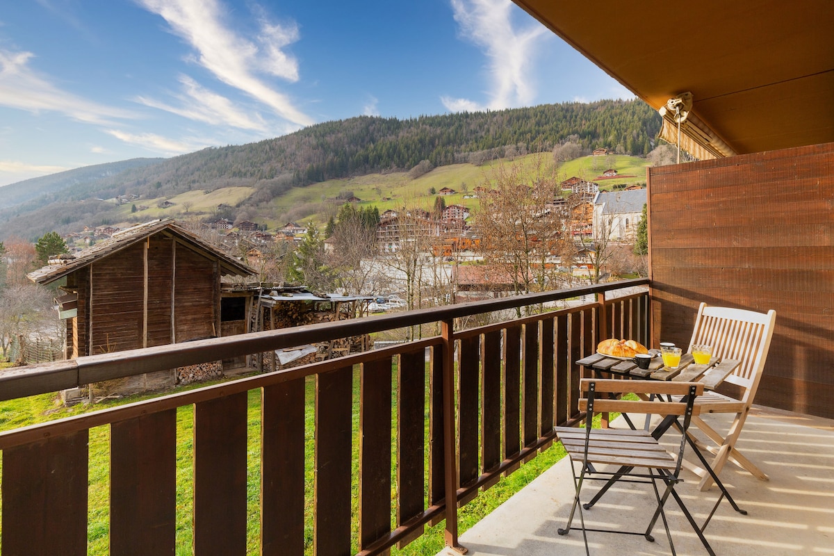 A furnished balcony features two chairs and a small table, set against a scenic view of rolling hills and distant buildings. The table is set with breakfast items, inviting a relaxing outdoor experience amidst nature.