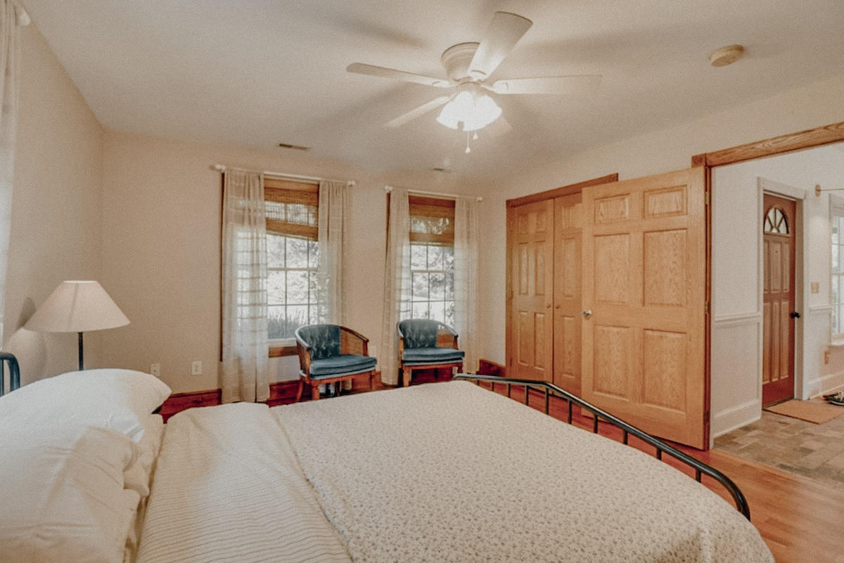 A spacious bedroom is furnished with a comfortable bed and twin armchairs near a window, offering natural light. Light-colored walls and hardwood floors create a bright atmosphere. A ceiling fan hangs above, and wooden doors provide easy access to the adjoining areas.