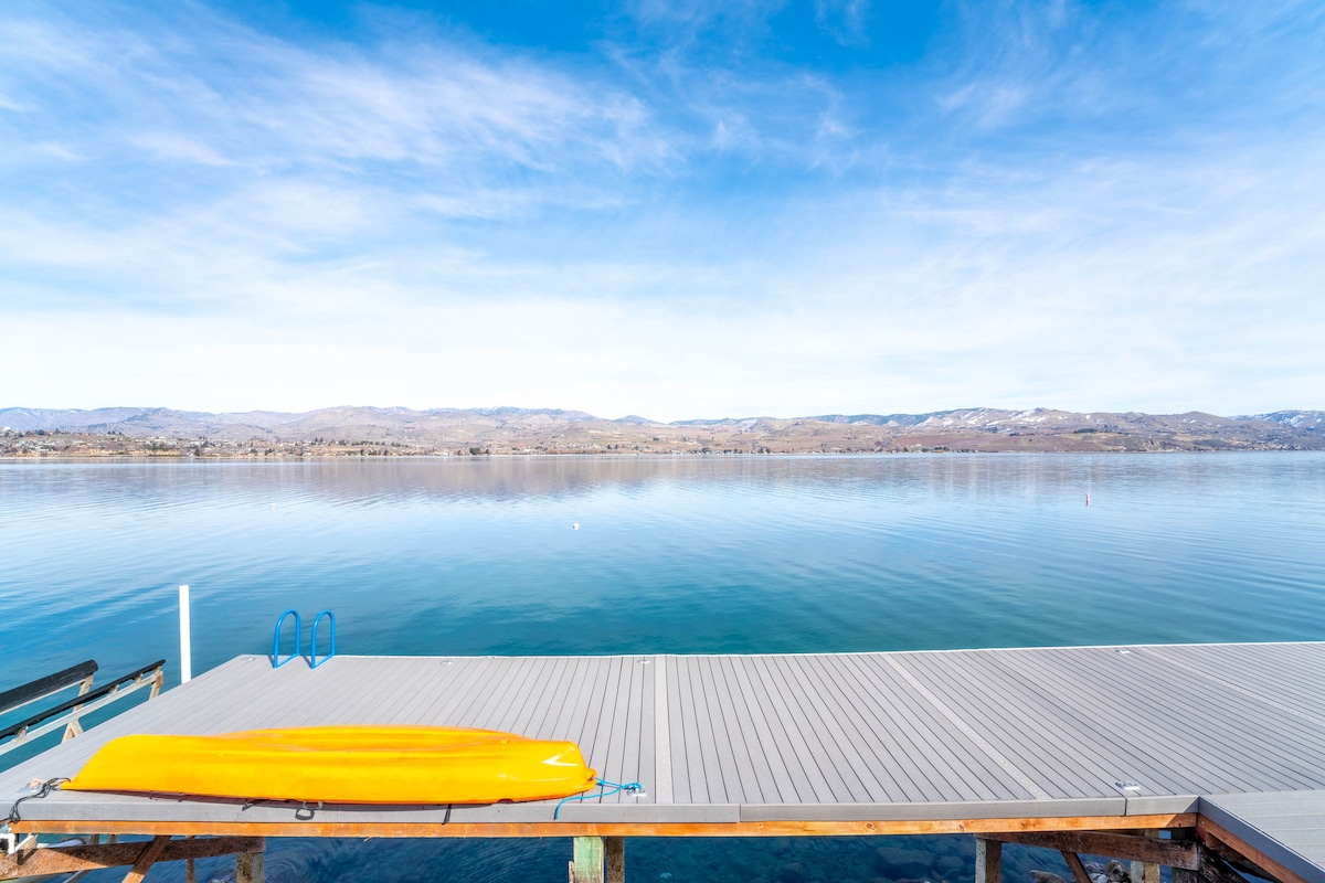 A shared dock extends into calm waters, showcasing a bright yellow kayak resting along the side. The serene lake surface reflects the surrounding landscape, with distant hills visible under a clear blue sky. Soft clouds drift above, enhancing the peaceful atmosphere.